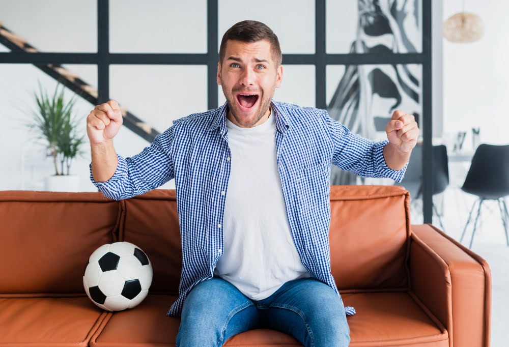 A man sitting on a sofa, smiling and raising his arms after winning a sports bet.