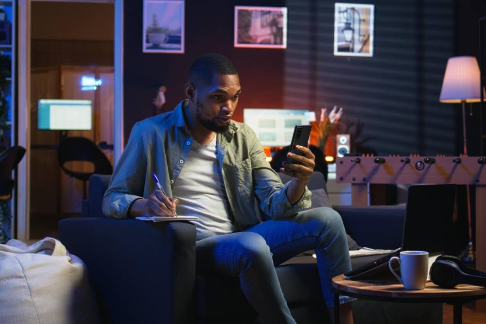A man sitting on a sofa looking at his phone to see when NFL player props come out.
