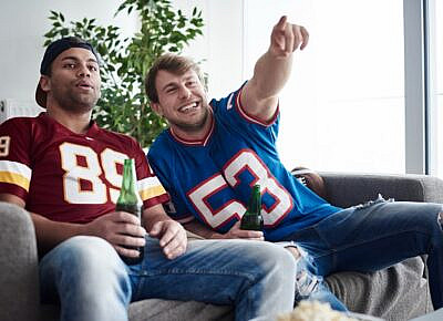 A couple of friends wearing NFL jerseys, cheering while watching a football game on TV.